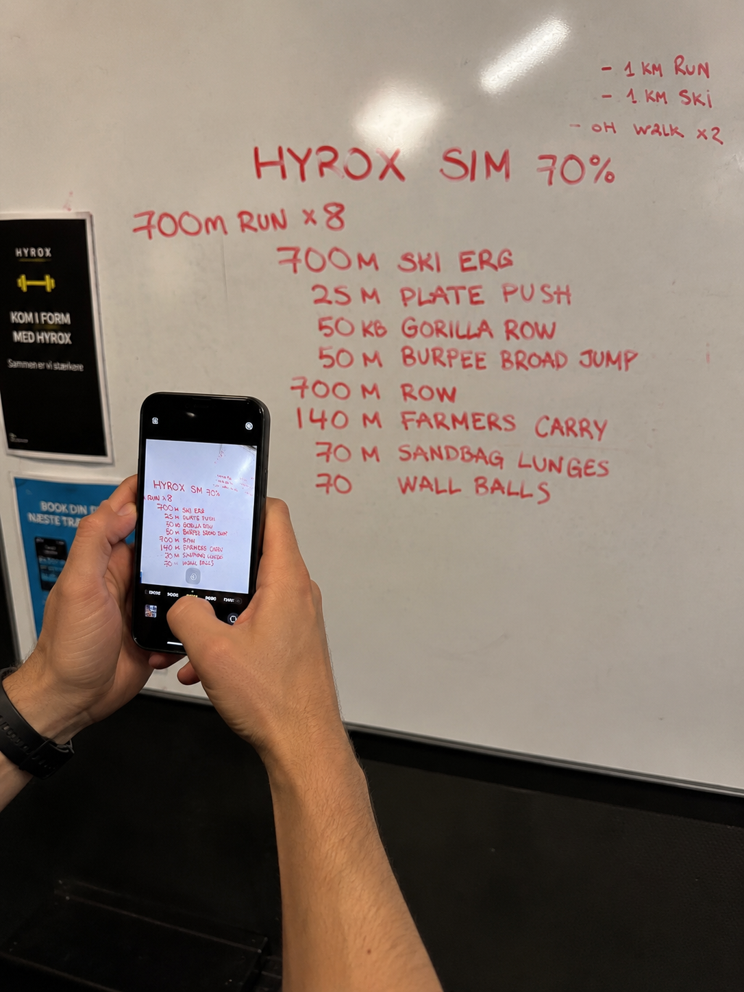 Athlete taking a phone photo of a HYROX workout whiteboard in a gym