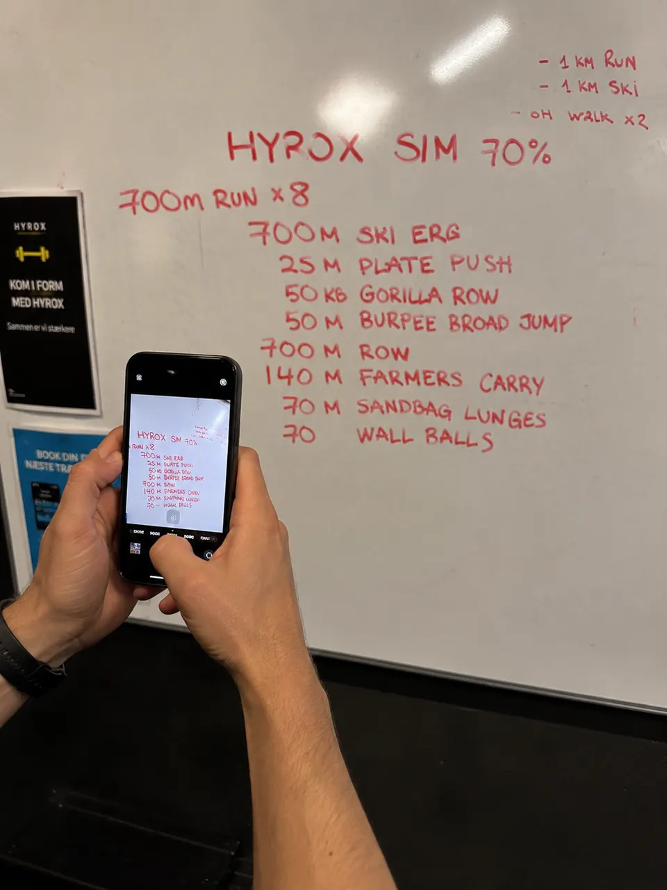 Athlete taking a phone photo of a HYROX workout whiteboard in a gym